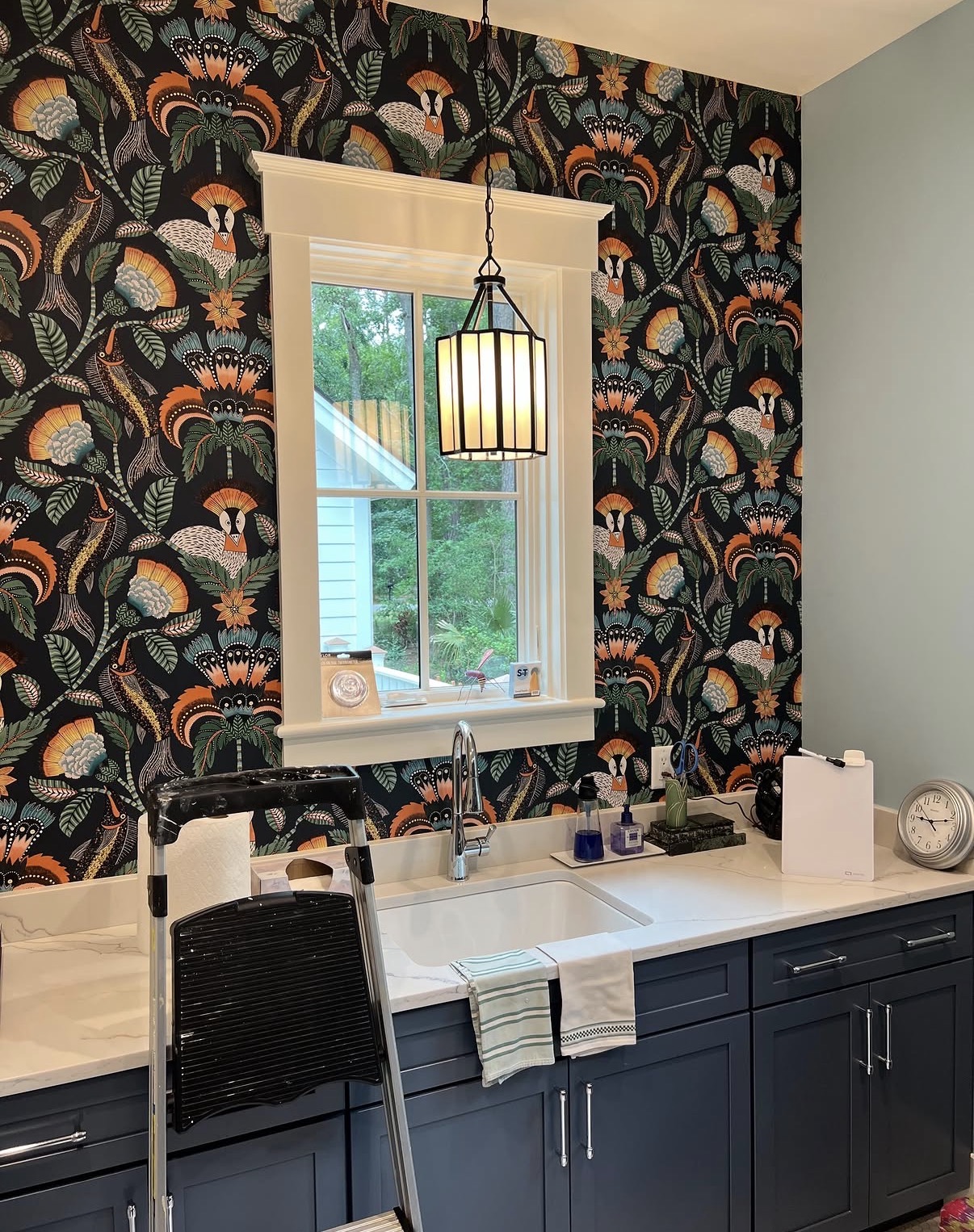 Folk-art bird and thistle wallpaper on black ground in a laundry room with navy cabinetry