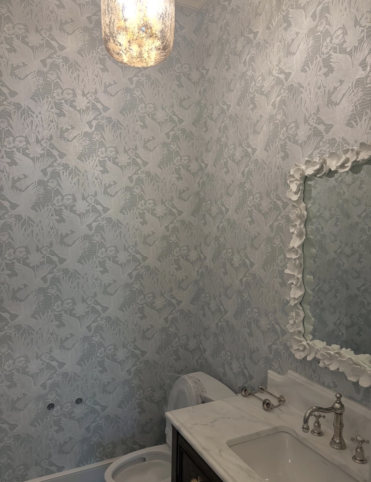 Pale silver-blue floral wallpaper in a powder room with sculptural plaster mirror